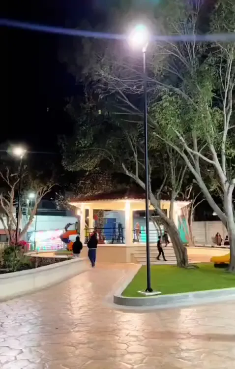 Parque municipal iluminado de noche con luminarias en postes, juegos infantiles y andadores